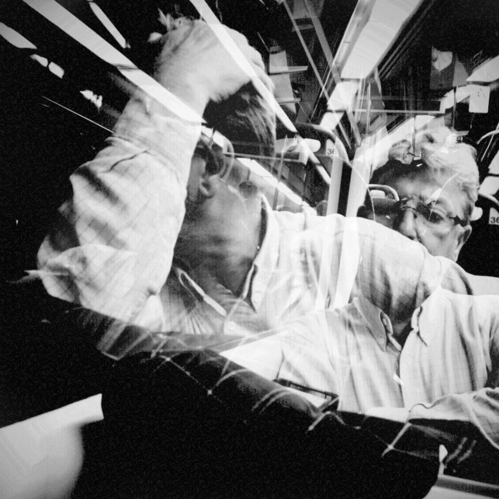 Black-and-white multiple-exposure photograph inside a train carriage showing a man raising his arm beneath bright ceiling lights, with several overlapping positions of his head and arm.
