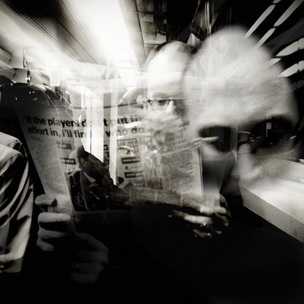 Black-and-white multiple-exposure image of a passenger reading a newspaper on a train, with overlapping views of their face and the newspaper layered across the frame.