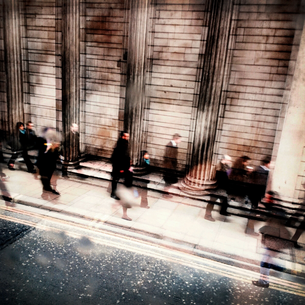 Blurred colour photograph of commuters walking along a pavement beside tall stone columns, their figures repeated along the pavement due to motion and multiple exposure.