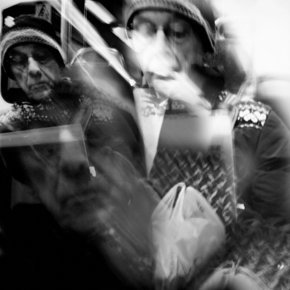 Black-and-white multiple-exposure photograph of a person wearing a knit hat and holding a plastic bag while seated on a train, with overlapping positions of their head and hands.
