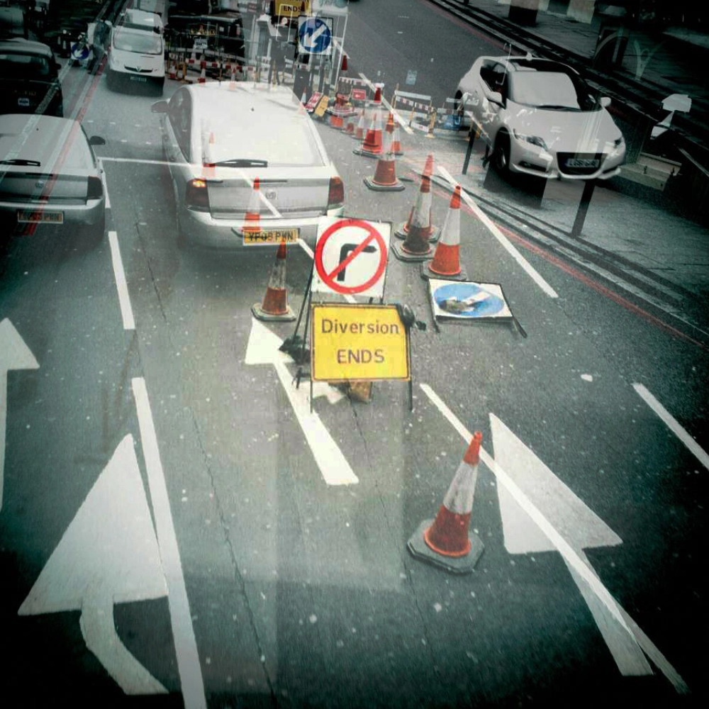 Colour photograph taken through a bus window showing roadworks with traffic cones and a sign reading “Diversion Ends,” with cars waiting in adjacent lanes.