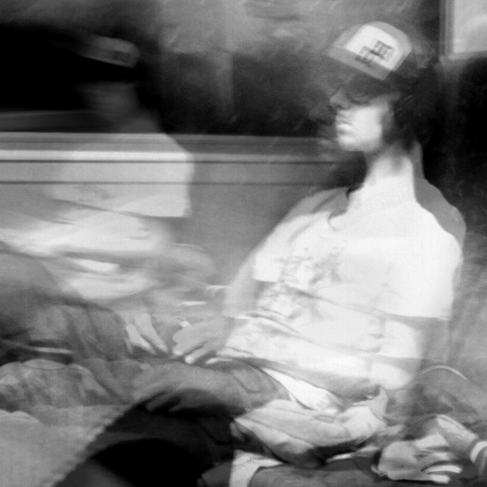 Black-and-white multiple-exposure photograph of a seated passenger wearing headphones and a baseball cap, with repeated positions of their torso and arms.