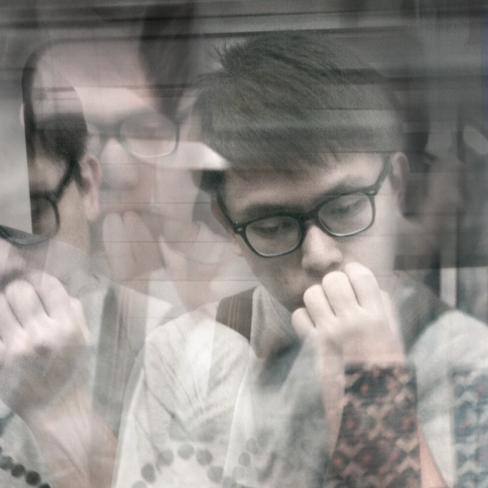 Black-and-white multiple-exposure portrait of a person with glasses resting their hand near their mouth, creating several overlapping views of the face.
