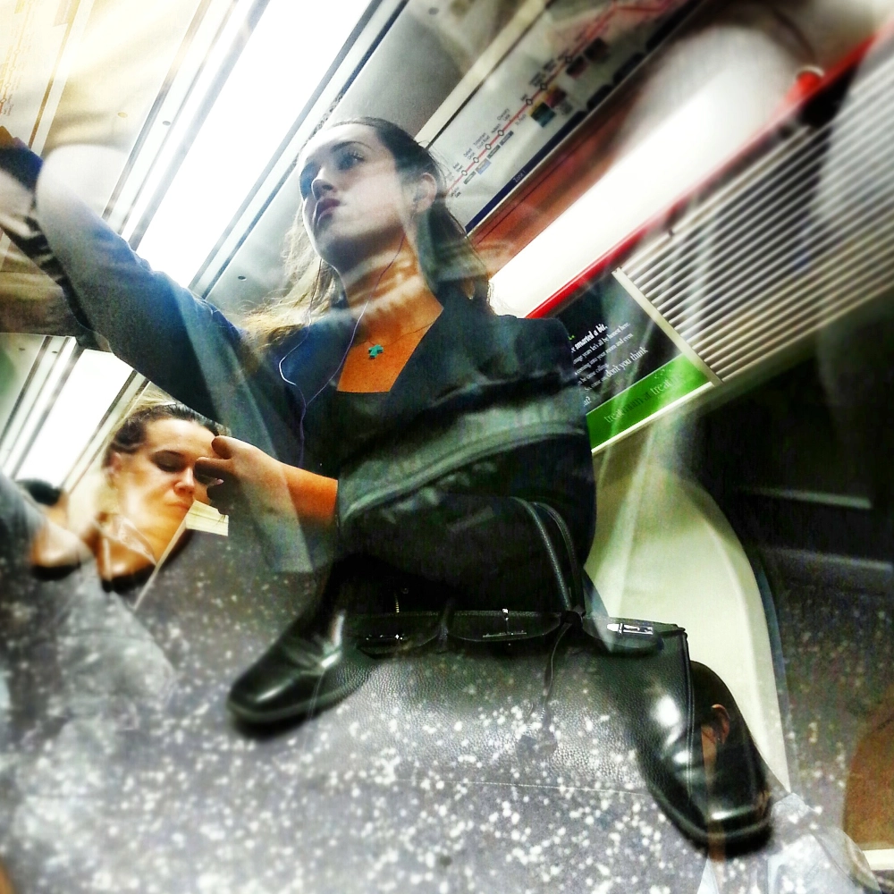 Colour multiple-exposure photograph inside a train carriage. A woman wearing earphones stands holding an overhead rail and looking forward. A second exposure overlays a pair of shiny black leather shoes across the lower part of the frame. Another passenger sits behind her with eyes closed. A transit map and carriage signage appear above.