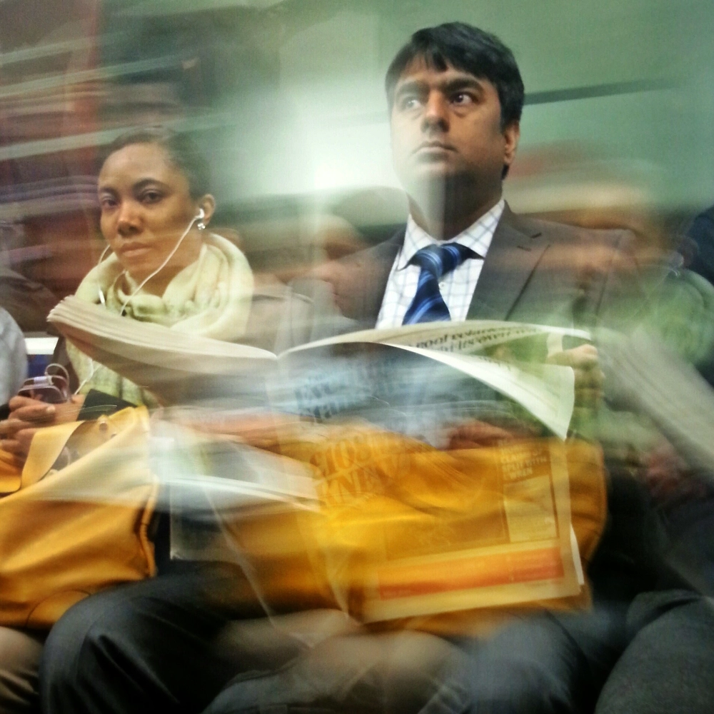 Colour photograph of two passengers seated on a train, a man reading a newspaper beside a woman wearing earphones, with slight motion blur across the frame.