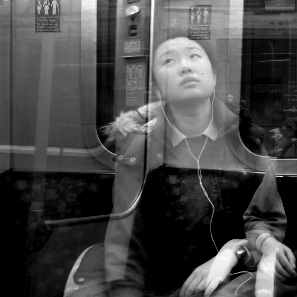 Black-and-white multiple-exposure photograph of a passenger wearing earphones seated by a train window, looking upward with overlapping positions of the head and shoulders.