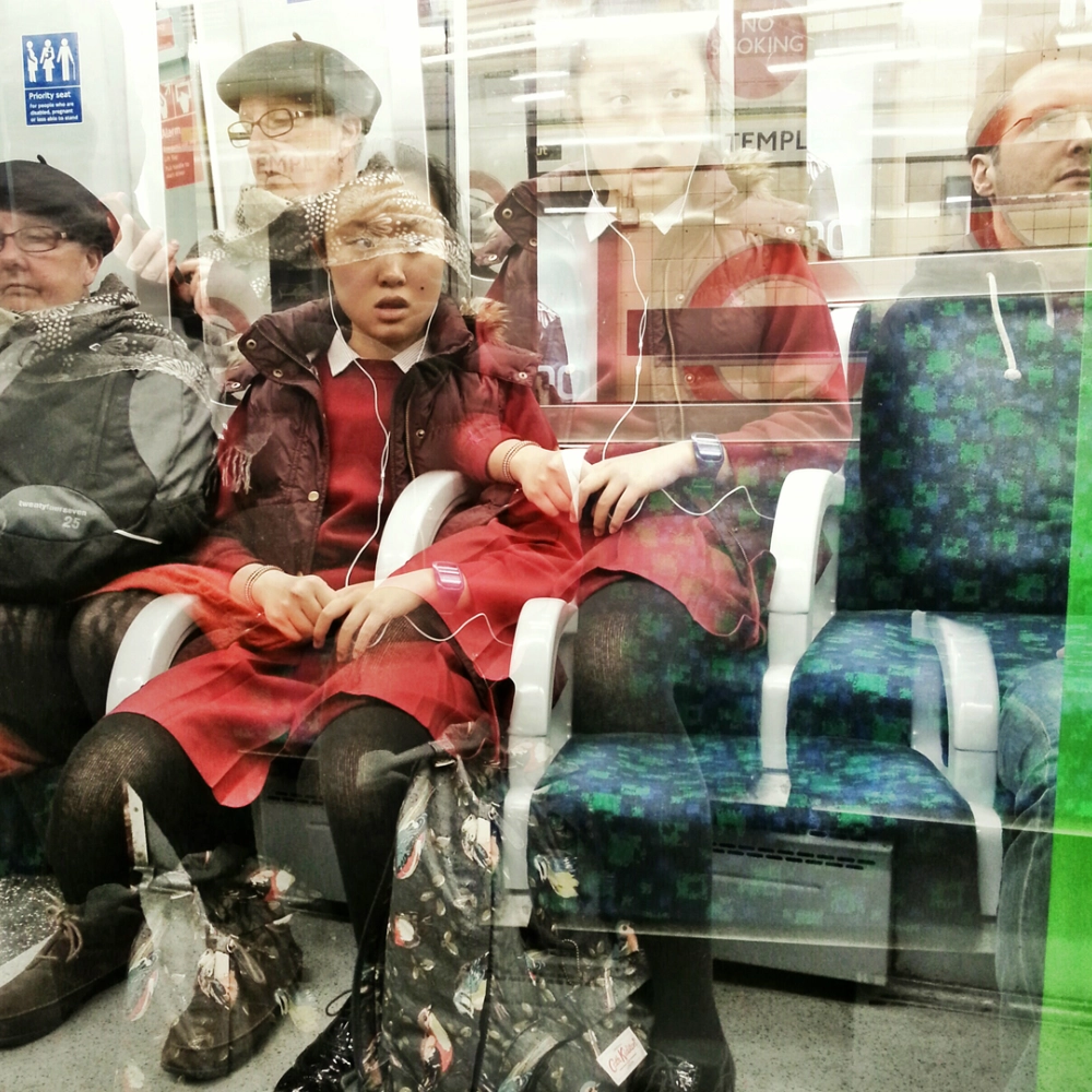 A multiple‑exposure photograph inside a subway carriage, layering repeated figures of seated passengers. A girl in a red jacket appears in several offset positions, with other riders and signage merging into the overlapping interior.