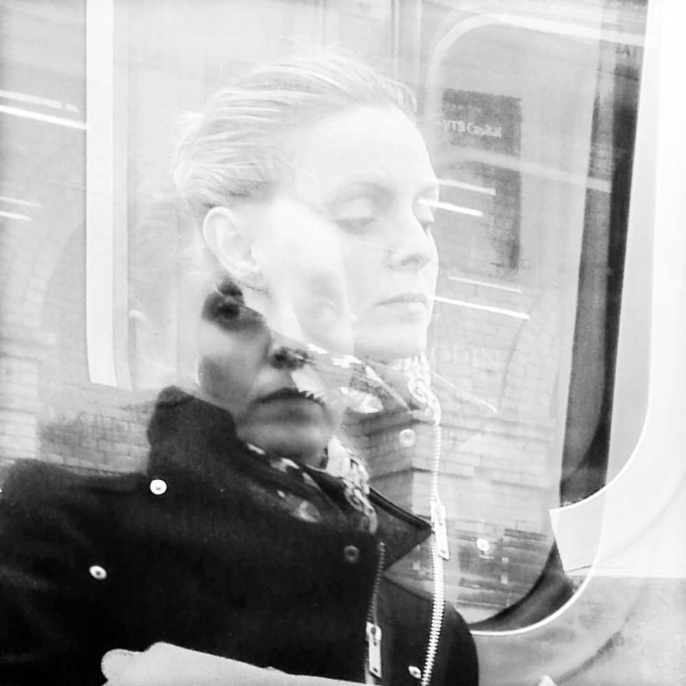A multiple‑exposure photograph on a subway carriage, showing the same person seated with their face appearing twice in slightly different positions, creating a layered doubled portrait against the interior and the passing brick wall.