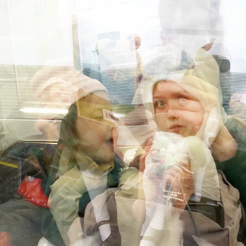 A multiple‑exposure photograph on a subway carriage, showing several overlapping images of two children in winter hats, their faces and gestures layered into a dense composite against the interior and window.