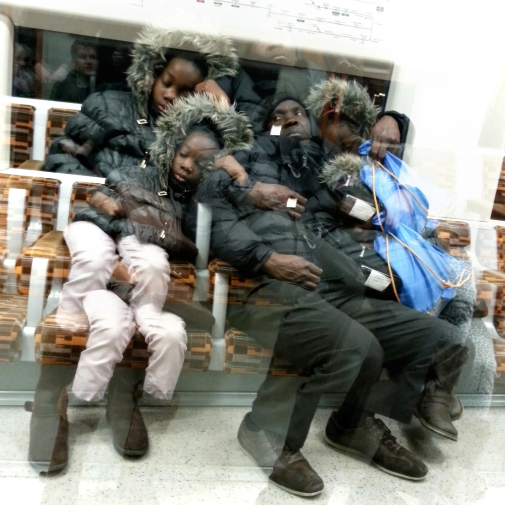 A multiple‑exposure photograph on a subway carriage, showing a father holding two children close, their duplicated bodies and winter jackets layered across the bench beneath a subway map.