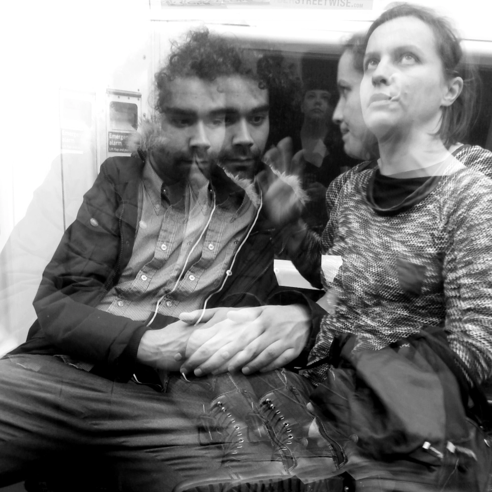 A multiple‑exposure photograph on a subway carriage, showing overlapping images of two people seated close together, their faces, jackets, boots, and leaning postures layered in several offset positions, with a faint figure behind them holding a mobile phone up in a gesture of taking a photo against the window and interior wall.