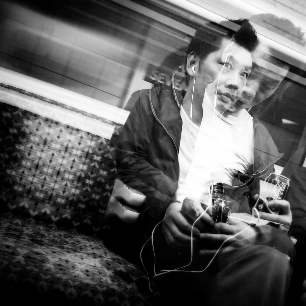 A multiple‑exposure photograph on a train, showing overlapping images of a person seated on a patterned bench, wearing a jacket and white shirt and holding a can of Coca‑Cola and a smartphone, their face, hands, and posture layered in several offset positions beneath the bright overhead light.
