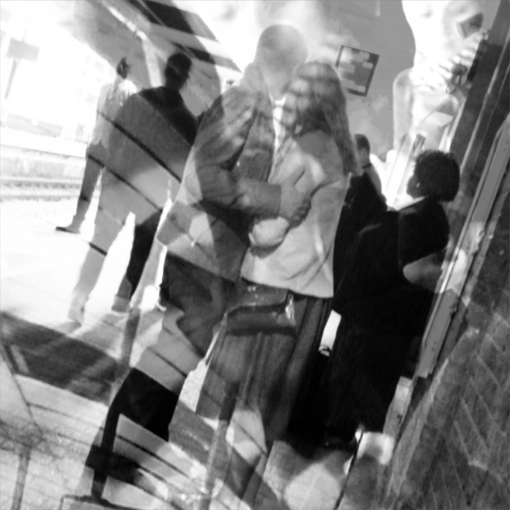 A multiple‑exposure photograph on a train platform, showing a couple embracing in the foreground, their faces, arms, and jackets clearly visible as other people around them appear in layered and offset positions across the platform.