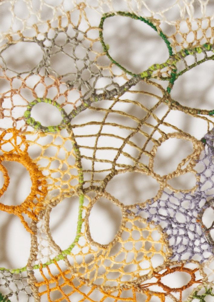 Detail showing a varied interior section with a large elongated form in olive-tan worked in a grid-like ladder stitch, surrounded by ring and oval outlines in green, lime, amber, and lavender. At lower right a dense lavender-purple filled patch is visible, and at lower left an open amber ring with a honeycomb interior. The surrounding ground is open cream net with scattered small loop forms.