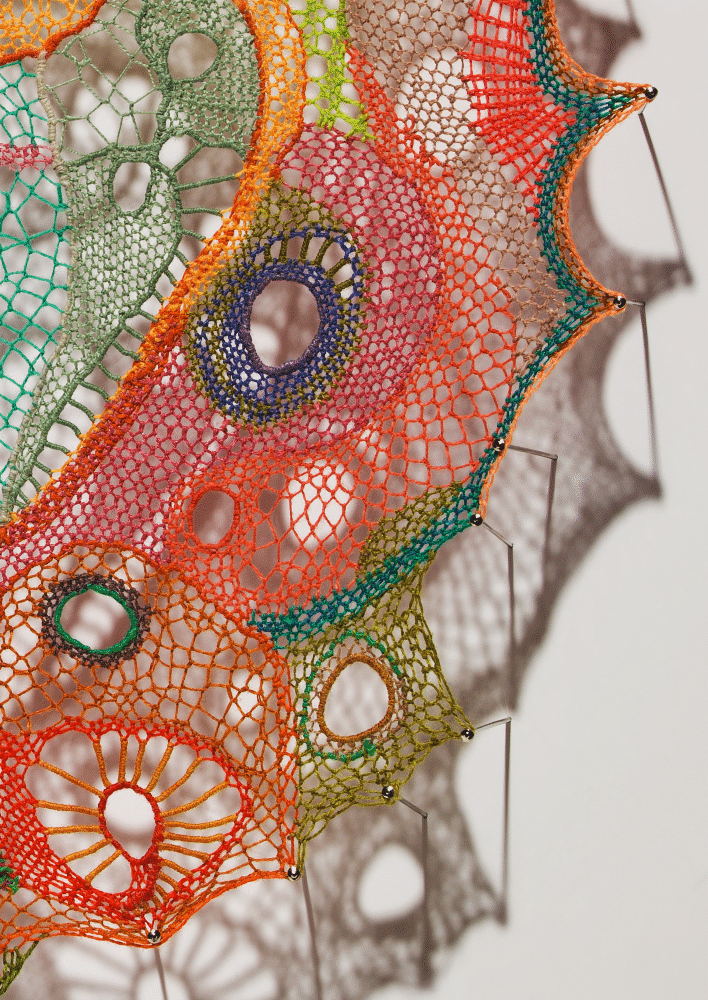 Detail against a white wall showing the lower-right quadrant with several sewing needles visible along the outer edge. The pointed outer border transitions from teal through orange and olive-green. Interior forms include a prominent ring with a blue-purple and orange surround at upper center, a small green-outlined ring at lower left, and a radial spoke motif in orange and yellow at lower center. A cluster of small multicolored rings in purple, green, and red appears at lower right. The piece's shadow is cast clearly on the wall.