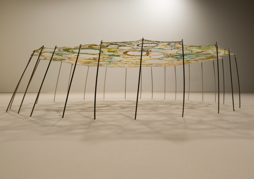 A low, straight-on view of "Metaphorical Cell No. 12: Pippa, Destroyer of Monsters" displayed horizontally, resting on a cream surface. The sewing needles pierce the outer edge of the circular embroidered textile and extend downward at outward-splaying angles, forming a ring of slender legs that elevate the piece above the surface like a low table or the body of a water-striding insect. The underside of the piece and the shadows cast by the needle legs are visible, with fine thread lines radiating outward across the surface below. Only a narrow strip of the embroidered textile edge is visible, showing hints of yellow-green and teal thread.